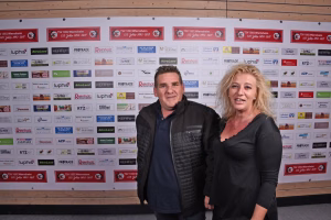 Zwei Personen vor einer Sponsorenwand mit zahlreichen Logos, stehen frontal in Outdoor-Kleidung und posieren für ein Gruppenfoto.