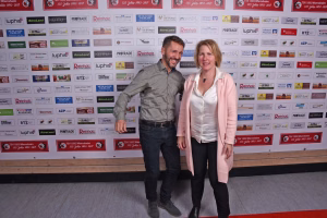 Zwei Personen posieren lächelnd vor einer Sponsorwand; stehende Portraitaufnahme für einen Event-/Pressekontext.