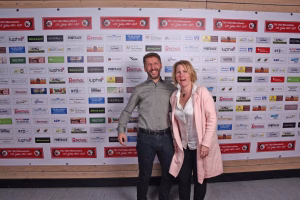Zwei Personen posieren nebeneinander vor einer Sponsorwand mit zahlreichen Logos; sie stehen frontal und wirken wie bei einem Eventfoto.