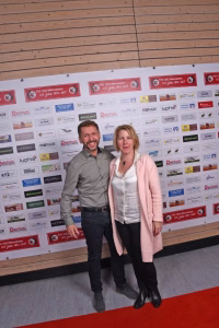Zwei Personen stehen vor einer bedruckten Sponsorenwand und posieren für ein Gruppenfoto, das den Veranstaltungsbesuch dokumentiert.
