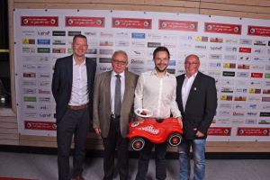 Vier Männer posieren vor einer Sponsorenwand; einer hält ein rotes Spielauto mit Fahrerhelm, die Aufnahme dokumentiert die Präsentation.