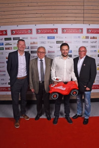 Vier Männer posieren auf rotem Teppich vor einer Sponsorenwand; ein rotes Spielzeugauto mit Reifenhalter wird gemeinsam präsentiert.