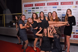 Gruppe feiernder Frauen posiert vor einer Sponsorenwand; mehrere schwingen einen goldenen Pokal als Siegergeste, Publikumseffekt bei Veranstaltung.