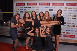 Gruppe junger Frauen posiert lächelnd vor einer Sponsorwand; Arme ausgestreckt, Blick in die Kamera, feierliches Gruppenfoto.