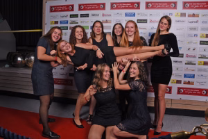 Gruppe von Frauen posiert gemeinsam für ein Gruppenfoto vor einer Sponsorwand, verschränkt Arme und jubelt, lachende Gesichter.