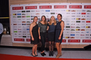 Vier Frauen stehen nebeneinander vor einer Sponsor-Wand und einem roten Teppich; sie posieren für ein Gruppenfoto.