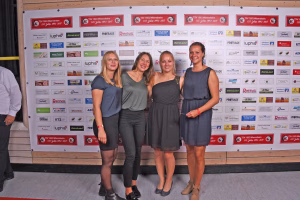 Drei lächelnde Frauen posieren vor einer Sponsorwand mit Vereinslogos; sie stehen im Vordergrund für ein Gruppenfoto bei einer Veranstaltung.