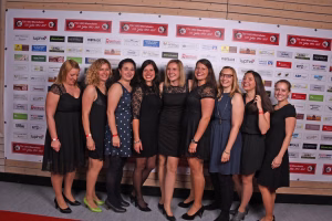 Gruppe von acht Frauen in Abendkleidung steht lächelnd vor einer Sponsorwand, dient als Gruppenfoto bei einer Veranstaltung.