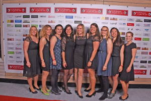 Gruppe von acht Frauen in dunkler Kleidung posiert lächelnd vor einer Sponsorwand mit vielen Logos für ein Mannschaftsfoto.