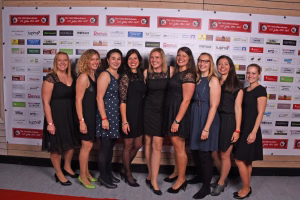 Gruppe von acht Frauen in festlichen Kleidern und Schuhen posiert lächelnd vor einer Sponsorwand als Porträt für Vereinsveranstaltung.
