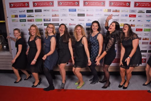 Gruppe von Frauen in Abendkleidung steht auf rotem Teppich vor einer Sponsorwand, lacht in die Kamera, für ein Eventfoto.