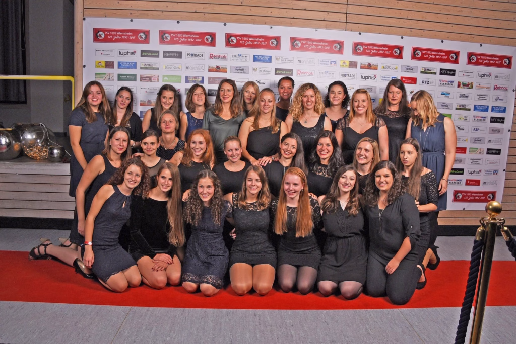 Gruppenfoto von mehreren Jugendlichen in dunkler Kleidung, die in zwei Reihen in einer Sporthalle vor einer Sponsorwand sitzen und lächeln.