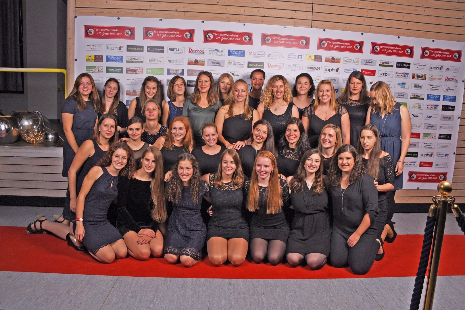 Gruppenfoto von mehreren Jugendlichen in dunkler Kleidung, die in zwei Reihen in einer Sporthalle vor einer Sponsorwand sitzen und lächeln.