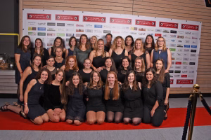 Gruppenfoto mehrerer Frauen vor einer Sponsorenwand und hölzernem Hintergrund, gemeinsam posierend; Zweck: Teampräsentation bei Veranstaltung.