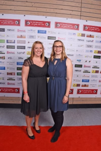 Zwei lächelnde Frauen posieren nebeneinander auf einem roten Teppich vor einer Sponsorwand mit Logos.