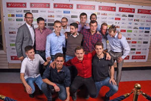 Gruppenfoto mehrerer Männer vor Sponsorenwand; einige stehen, andere sitzen und posieren lächelnd, Zweck: gemeinsamer Anlass/Teampräsentation.