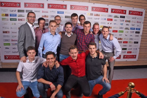 Teamfoto mehrerer Männer vor einer Sponsorwand, posierend in zwei Reihen; zentrale Gruppe im Vordergrund mit Arm um die Schultern.
