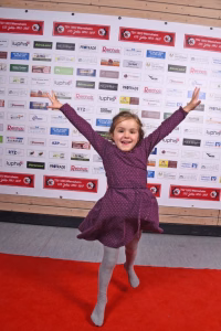 Mädchen in lila Kleid mit ausgestreckten Armen und hochgehobenem Bein auf rotem Teppich vor Sponsorwand; zeigt fröhliche Pose im Eventbereich.