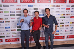 Drei Männer stehen auf einer Bühne vor Sponsor-Wand, halten Biergläser in der Hand und lächeln. Hintergrund mit Firmenlogos und Jubiläumsplakaten.