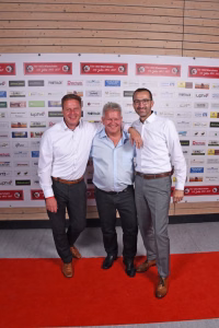 Drei Männer posieren lächelnd auf rotem Teppich vor einer bedruckten Sponsorwand; sie stehen eng beieinander und winkeln/armen über die Schulter.