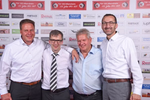 Vier Männer in weißen Hemden posieren lächelnd nebeneinander vor einer Sponsorwand; Personengruppe bei Veranstaltung/Teamfoto.