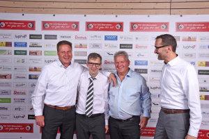 Drei Männer in Hemden posieren vor Sponsorwand mit vielen Logos; einer schaut zur Seite, die anderen lächeln, Hintergrund dient als Kulisse.