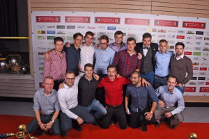 Gruppe von Männern posiert eng zusammen vor Sponsorwand bei Veranstaltung; Hände zur Seite, einige hocken, Fokus auf gemeinsames Mannschaftsfoto.