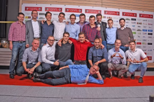 Gruppe von Personen posiert gemeinsam auf rotem Teppich vor einer Wand mit Vereinsplakaten, zeigt freundliche Gesten und lächelndes Gruppenbild.