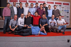 Gruppenfoto von Männern bei einer Veranstaltung; sie posieren in mehreren Reihen vor einer Sponsorwand, teils lächelnd, Arm um Schulter.