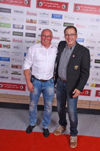 Zwei lächelnde Männer stehen nebeneinander vor einer Sponsorwand, posieren bei einem Event mit runder Themen-Dekoration im Hintergrund.