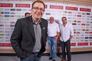 Mann im Anzug posiert vor einer Sponsorenwand mit mehreren Logos, im Hintergrund zwei weitere Personen; Bild dokumentiert ein Team- oder Veranstaltungsfoto.
