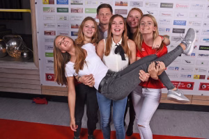 Gruppe junger Leute posiert vor einer Sponsorwand; eine Person lehnt lachend mit großem Jeansbein im Arm, alle schauen in die Kamera.