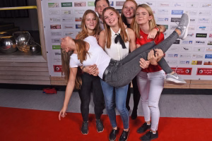 Mehrere Jugendliche posieren für ein Gruppenfoto, im Vordergrund schieben sie sich spielerisch auf einer großen grauen Sportstange vor einer Themenwand.