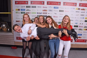 Gruppe junger Menschen posiert vor einer Sponsorwand und hält gemeinsam eine große, gepolsterte Staffelei—feierliche Siegespose.