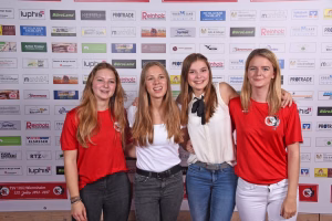 Vier junge Frauen posieren eng beieinander vor einer Sponsorwand, lächeln in die Kamera, wirken wie bei einem Foto für ein Event.