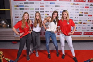 Vier junge Frauen posieren dicht beieinander vor einer Sponsorwand, lächeln in die Kamera; abgestimmte Freizeitkleidung als Gruppenfoto auf rotem Teppich.