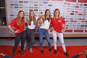 Vier junge Frauen stehen posierend vor einer Sponsorwand, drei in Rot-Weiß, eine links in Rot-Grau, auf rotem Teppich.