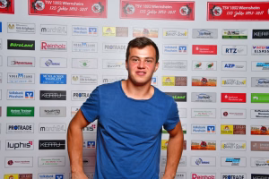 Mann im blauen T‑Shirt steht vor einer Wand mit vielen Logos und tritt als Interview-/Fotomotiv hervor.