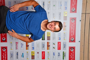 Mann in blauem T‑Shirt reicht mit erhobenen Armen einen Stoffbezug über die Schulter; Hintergrund mit vielen gedruckten Logos und Plakaten.