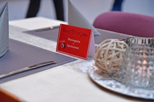 Rote Programmkarte mit Text „Ehrengäste + Sponsoren“ auf grauem Buffet, daneben gestreute Bestecke und dekorative Kerze.