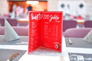 Rote zweiflügelige Getränkekarte mit Aufschrift und Zutatenliste, im Vordergrund vor einem gedeckten Bankettbereich.