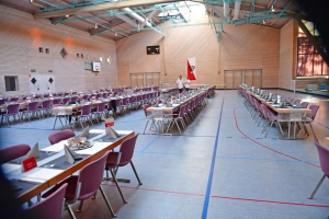 Mehrere gedeckte Reihen an Tischplätzen mit lila Stühlen in einer Sporthalle, perspektivisch für ein Event oder Catering angeordnet.