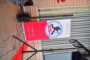 Rot-weißer Werbebanner mit Hundekopf-Logo an einer Wand, daneben Kabel und ein langes Metallgestell; dient zur Information.