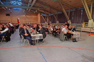 Konferenzteilnehmende sitzen an langen Tischen in einer Turnhalle, vorbereitet für ein offizielles Meeting mit Mikrofonen und Beleuchtung.