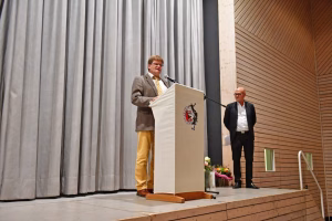Redner hinter einem Pult mit Mikrofon auf einer Bühne, daneben ein zweiter Mann, im Hintergrund Vorhänge und Holzpanelen.