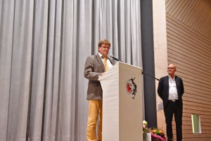 Redner steht an einem Podium mit Mikrofon vor Vorhang und Holzwand, hält eine Ansprache bei einer Veranstaltung.