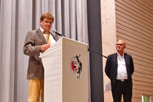 Redner am Podium mit Mikrofon und Emblem; im Hintergrund steht eine weitere Person neben einer Steinverkleidung und Vorhang.