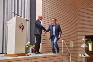 Zwei Männer auf einer Bühne geben sich die Hand; Holzfassade im Hintergrund, Mikrofonanlage und Deko-Blumen an der Seite.