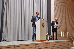 Mann hält einen Vortrag am Rednerpult mit Mikrofon; im Hintergrund liest eine zweite Person von Papier, vor Bühnenvorhang und Holzpaneelwand.