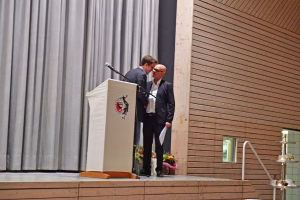 Zwei Redner am Podium küssen sich, während ein Mikrofon auf der Bühne steht; Blumenarrangement und Vorhang zeigen den Veranstaltungsrahmen.
